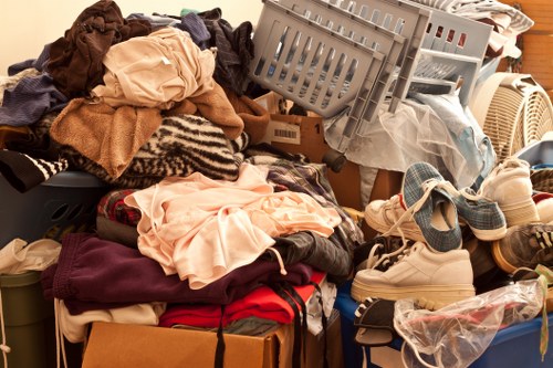 A large pile of assorted household rubbish and textiles, including folded and crumpled clothing in various colours such as beige, red, and black, is seen on a flat surface like a driveway or garage floor. Among the textiles are cardboard boxes filled with more items, alongside plastic containers and laundry baskets positioned at different angles. Shoes, including a pair of sneakers, are scattered on the pile or nearby, with some partly covered by other rubbish. In the background, a white plastic laundry basket and a beige appliance, possibly a heater or fan, are partially visible. The overall scene depicts a cluttered collection of disused household items, typical of a clearance or rubbish removal process, which a local service like House Clearance Lee could help manage in the Lee and nearby areas within the post code region.