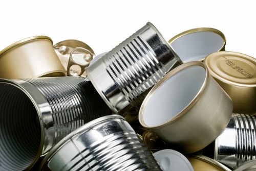 A collection of various empty tin cans, including some with metallic silver finishes and others with beige or gold-colored coatings, stacked and scattered on a flat surface. The cans vary in size and shape, with some lying on their sides and others upright, displaying smooth, cylindrical bodies with ridged or smooth sides. The background is neutral and blurred, focusing solely on the cans, which appear to be used for food storage or packaging. This assortment of discarded metal containers is typical of waste material handled by rubbish removal services, such as those provided by House Clearance Lee, serving areas including Lewisham and nearby towns. The textures of the cans are shiny and reflective, with some showing faint dents or scratches from previous use. The image captures the environmental context of waste disposal, highlighting the importance of proper rubbish collection and recycling processes in urban and suburban environments.